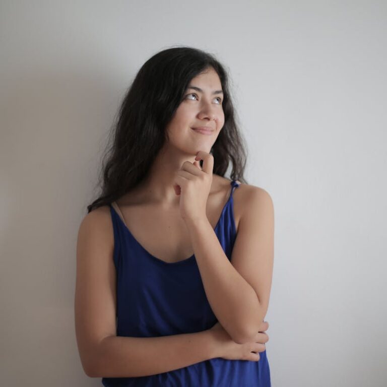 A pensive woman in a blue tank top stands against a plain white wall, smiling gently.